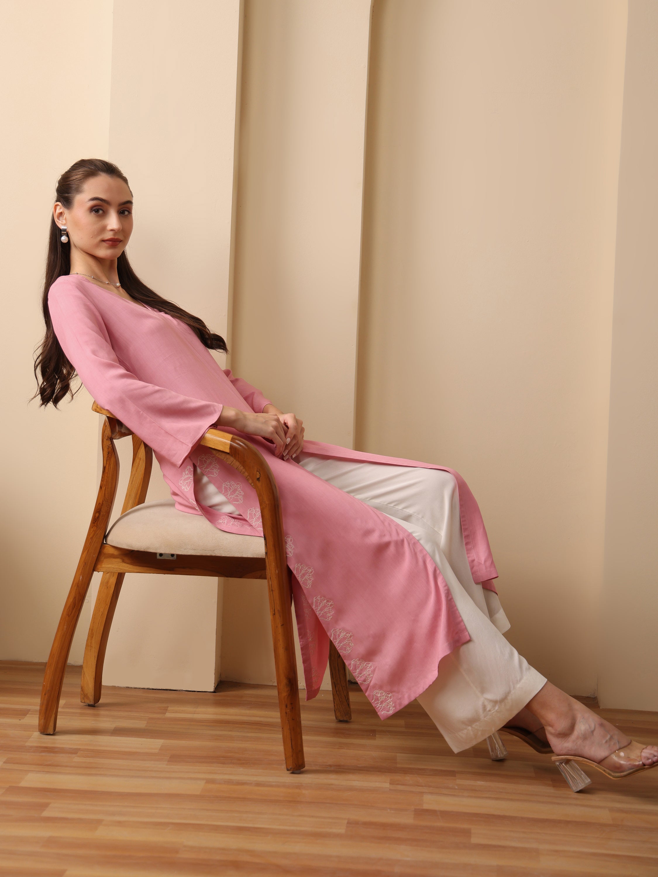 Woman wearing a pink long kurta with white embroidered palazzo pants sitting on a chair.